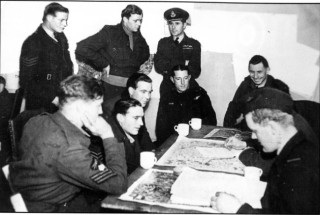 069_Ch_9_photo1 W/Cdr Comm talks to C/Capt Swain whilst the crew of P/0 C.5. Mant RAAF are given their post-raid debriefing by an Intelligence Officer. The Mant crew were shot down on the night of 11/12th March 1943 after attacking Stuttgart | ©️Vincent Holyoak