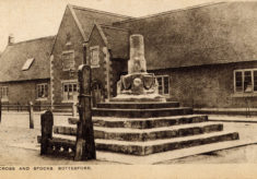 Postcard of the Market Cross and Stocks, with the village school behind.