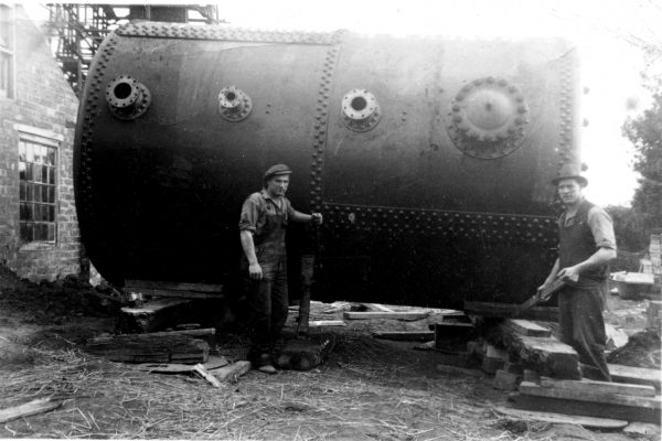 Men standing by a large cylindrical boiler.