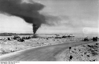 Image from the German Federal Archives shows: “The road from Bardia to Tobruk on 21 June 1942 with British prisoners of war on the left, sunken ships in the harbour and smoke over the port.” | Wikipedia