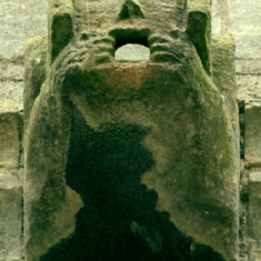 P1050516_05 One of the gargoyles on the tower, with mouths for water spouts: a face set in box-like head-dress, its hands are pulling its mouth wide open.