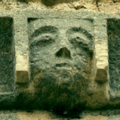 P1050518_06 One of the six small grotesque faces carved in the stones forming the frieze below the parapet of the tower. In this one, the face is set in a square box-like structure which may be a style of head-dress.