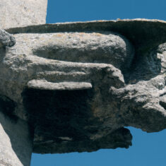 Two figures jut out one above the other on the eastern corner of the southern side of the clerestory. This, the lower one, is a winged creature with scaly legs ending in paws or clawed feet, and a human face. | Neil Fortey