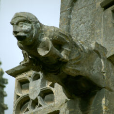 P1070364_06 On the south transept - The “bellman” or “tollman”: the western of the gargoyles on the parapet of the south transept. This almost life-size figure was carved in great detail, with clappers in his bells and buttons on his coat. His robust, jovial appearance suggests perhaps an invitation to the market rather than a religious intent. | Neil Fortey