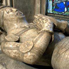 Sir John de Roos, 8th Baron Roos, d.1421. North of the altar is the alabaster tomb and effigy, brought from Belvoir Priory, of John, son of Sir William de Roos, who fought at Agincourt but lost his life at the decisive French victory at Baugé in 1421. Lord John also wears a conical bascinet with an orle, his head resting on a tilt-heaume which once bore the de Roos peacock crest. The alabaster is marked by graffiti, some of considerable age. | Neil Fortey