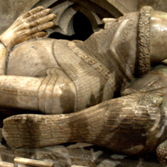 Tomb and Monument of Sir William de Roos, 7th Baron Roos, d. 1414. South of the altar is the alabaster tomb and effigy of William de Roos, a supporter of Henry Bolingbroke (Henry IV). This tomb together with that of his eldest son John (below) was brought from Belvoir Priory on the Dissolution of the Monasteries. The effigy and decorated base chest suggest the work of the Chellaston school of sculptors in Derbyshire. The armour is a mixture of mail and plate. A conical bascinet helmet with a jewelled margin is worn while under the head is a tilting-heaume and a peacock . On the front of the helmet is “IHC NASARE” (Jesus of Nazareth) and from the helmet hangs a tippet of chain mail over which is a decorative SS collar (SS stands for ‘Souvenir Sovereign’ - remember your king, a Lancastrian emblem). The insignia of the Garter is worn on the left leg. Behind the tomb is an Early English piscina. | Neil Fortey