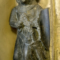 Sir Robert de Roos, 1st Baron Roos, d.1285. This Purbeck Marble effigy on the north wall of the Chancel was brought from Croxton Abbey during the Dissolution of the Monastries (1536-1541). It depicts a late 13th century knight wearing a hooded hauberk of chain mail under a loose sleeveless surcoat, mail gauntlets, a sword and belt on the left side together with a shield. It is accompanied by a plaque known as the ’Heart Stone’, also from Croxton Abbey, whose inscription records the burial of the heart of Robert de Roos at Croxton. In translation, the inscription reads: ‘Here lies the heart of Lord Robert de Roos, whose body is buried at Kirkham, who died on 13th May AD 1285. Isabella Lady de Roos, wife of the said Robert de Roos, was buried at New Place (Newstead) near Stamford, who died AD 1301”. She was the last of the Albini line at Belvoir. Her intricately carved sandstone effigy can be seen in St Mary’s, Orston, where it was moved to from Newstead (near Stamford). | Neil Fortey