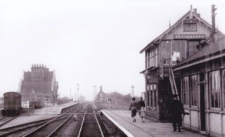 Picture 14#2 Looking westwards, showing the passenger crossing at rail-level between the staggered platforms, the signal box and waiting room on the up platform, the main station buildings on the down. In the distance are the station crossing-keeper’s house and further on the crossing-keeper’s house on Normanton Lane. | From an old postcard, possibly 1950s. Bottesford Local History Archive
